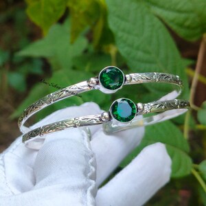 May include: Two silver bangles with a floral pattern and a large, round, emerald-green gemstone. The bangles are displayed on a white surface, with green foliage in the background. The text "New Silver Generation" is visible.