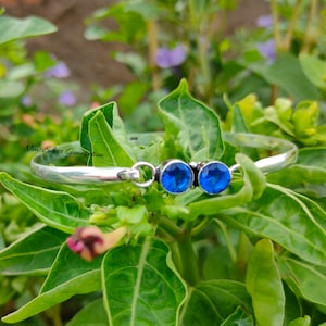 以下が含まれることがあります： 2つの青い宝石のアクセントが付いたシルバーのバングルブレスレット。ブレスレットは開いており、ヒンジ付きの留め具が付いています。