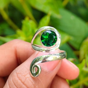 May include: A silver ring with a large, round, faceted emerald green gemstone. The ring has a swirling, open design with the text "New Silver Generation" on the band. The ring is held by a person.