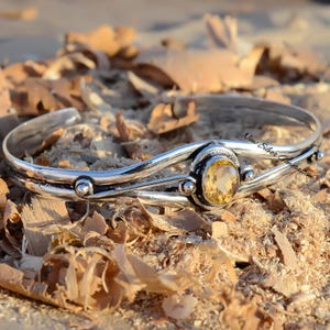 Op de afbeelding: Een zilveren armband met een enkele, ronde, gele citrien edelsteen gezet in een bezel setting. De armband heeft een golvend ontwerp en is versierd met kleine zilveren kralen.