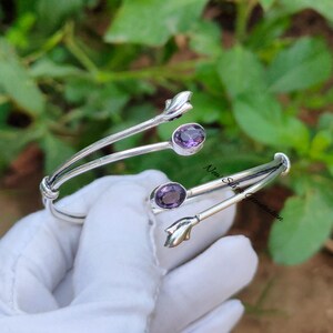 May include: A silver cuff bracelet featuring two oval, purple gemstones. The bracelet has a delicate, open design with floral accents. The text "New Silver Generation" is visible on the bracelet.