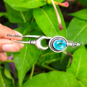 May include: Silver bangle bracelet with a crescent moon design and a turquoise blue gemstone. The bracelet is open and has a simple design.
