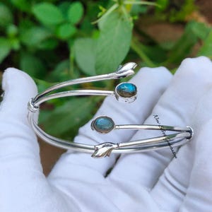 Op de afbeelding: Een zilverkleurige manchetarmband met twee ovale, blauwgroene edelstenen. De armband heeft een uniek ontwerp met bladachtige accenten en is open. De tekst "New Silver Generation" is zichtbaar.