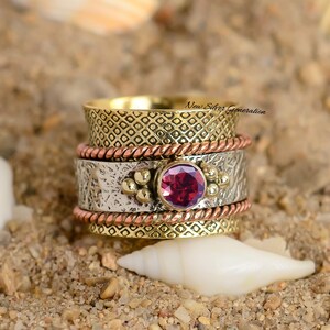 May include: A multi-layered ring with a large, round, red gemstone at its center. The ring features bands of gold, silver, and copper, with textured and patterned designs. The ring is set against a sandy background with a seashell.