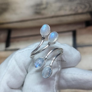 Può includere: Un anello argentato con quattro cabochon ovali di pietra di luna. L'anello ha un design a bypass, con ogni pietra incastonata su una fascia curva. Le pietre di luna hanno una base bianca lattea con iridescenza blu.