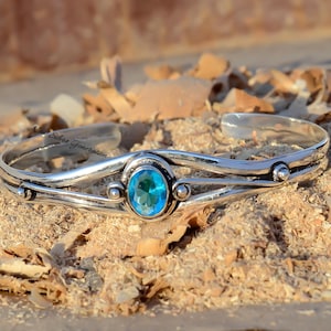 May include: A silver cuff bracelet with a blue gemstone set in the center. The bracelet has three bands and is decorated with small silver beads.