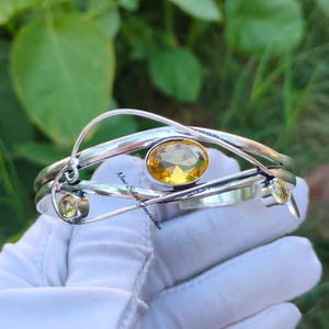 Könnte beinhalten: Ein silbernes Armreif-Armband mit einem gelben Edelstein und zwei kleineren gelben Edelsteinen. Das Armband hat ein verdrehtes Design und ist aus Sterlingsilber gefertigt.