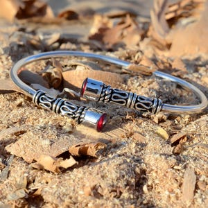 May include: Silver bangle bracelet with two red gemstone accents. The bracelet has an intricate design with a braided pattern.