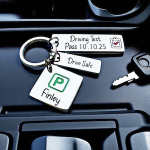 May include: A silver keychain with three rectangular tags and a square tag. The tags read "Driving Test Pass 10.10.25", "Drive Safe", and "Finley" with a green "P" symbol. A car key and ignition are also visible.
