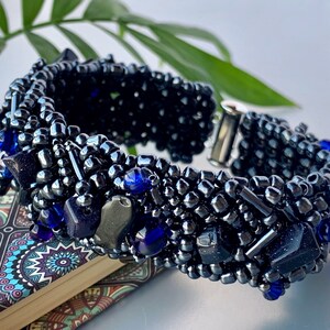 Puede incluir: Un brazalete de cuentas negras con cuentas de vidrio azules y un cierre de metal plateado. El brazalete está hecho con una variedad de cuentas de diferentes tamaños y tiene un aspecto texturizado único.