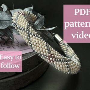 Puede incluir: Un brazalete de cuentas blancas, plateadas y rosas con cierre de broche. El brazalete está sobre una superficie gris con una tarjeta rosa que dice "Fácil de seguir" y una tarjeta rosa que dice "Patrón PDF + video".