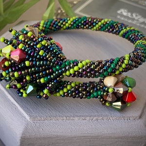 Könnte beinhalten: Ein Perlenarmband mit Spiralmuster. Das Armband hat eine Mischung aus grünen, gelben und irisierenden Perlen. Dekorative Cluster aus facettierten Perlen in Rot, Grün und Beige sorgen für Details. Das Armband wird auf einer weißen Oberfläche präsentiert.