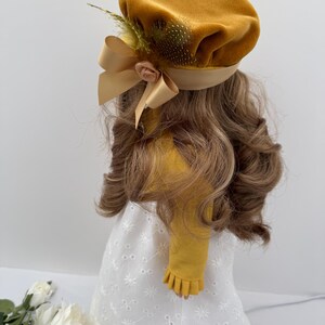 May include: Mustard yellow velvet beret with a satin ribbon bow and a small fabric rose. The beret is adorned with a feather accent. The beret is part of a doll's outfit, which includes a white eyelet dress and long, wavy brown hair.