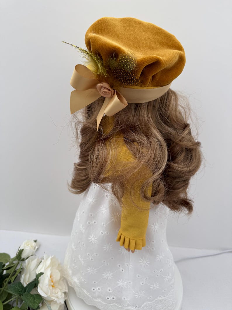 May include: Mustard yellow velvet beret with a satin ribbon bow and a small fabric rose. The beret is adorned with a feather accent. The beret is part of a doll's outfit, which includes a white eyelet dress and long, wavy brown hair.