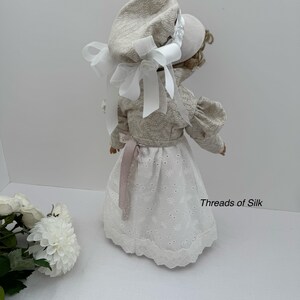 Puede incluir: Una mu&ntilde;eca con un gorro y chaqueta color crema con lazos blancos. La mu&ntilde;eca tiene una falda de encaje de ojal blanco y una cinta rosa. El texto "Threads of Silk" es visible en el lado derecho de la imagen.