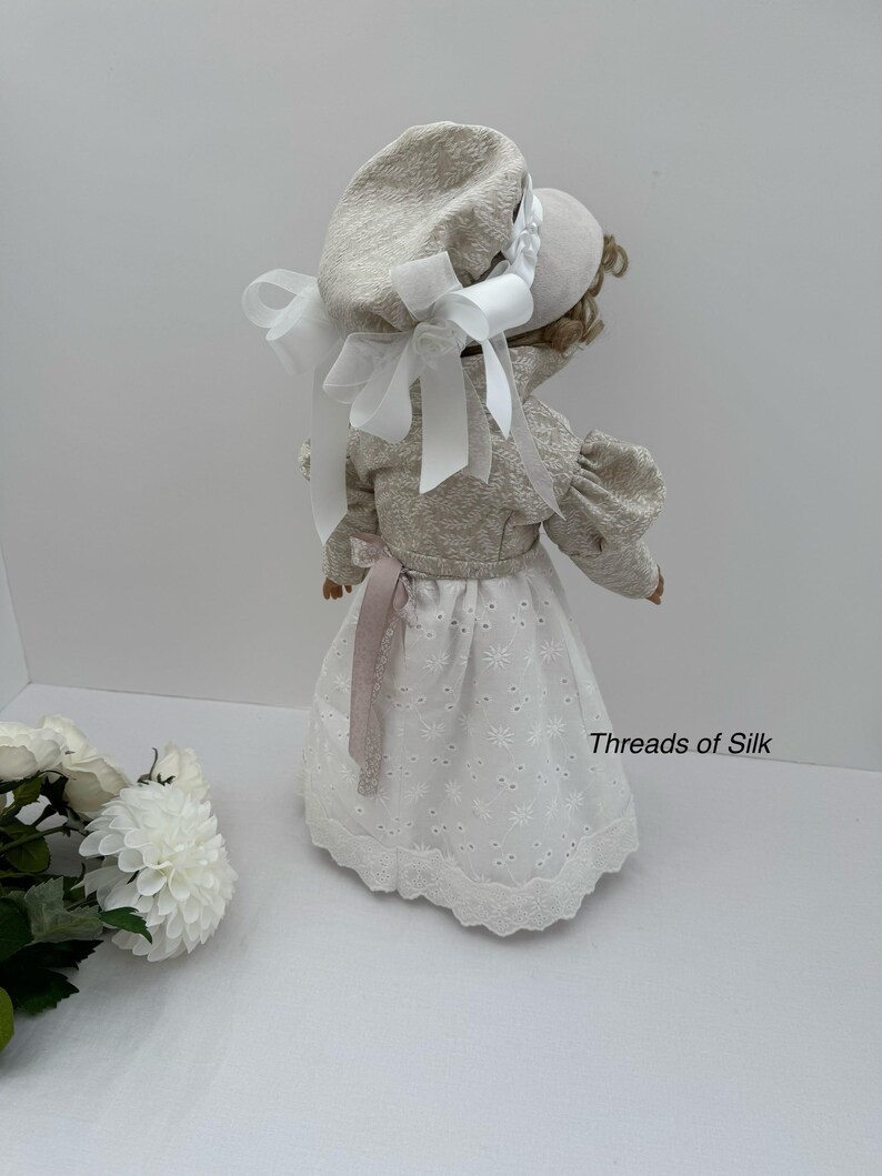 Puede incluir: Una mu&ntilde;eca con un gorro y chaqueta color crema con lazos blancos. La mu&ntilde;eca tiene una falda de encaje de ojal blanco y una cinta rosa. El texto "Threads of Silk" es visible en el lado derecho de la imagen.