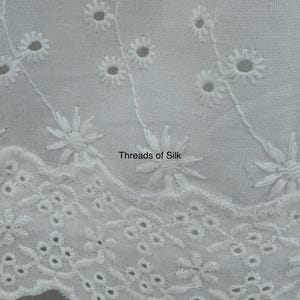 Puede incluir: Primer plano de tela calada blanca con dise&ntilde;os florales y geom&eacute;tricos recortados. La tela presenta flores bordadas en forma de margarita y el texto "Threads of Silk". El borde festoneado a&ntilde;ade un toque decorativo a este textil.