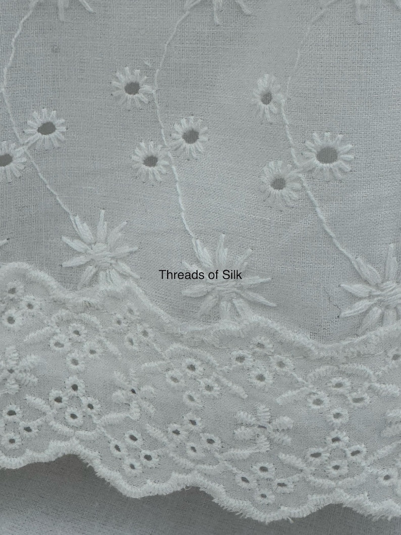 Puede incluir: Primer plano de tela calada blanca con dise&ntilde;os florales y geom&eacute;tricos recortados. La tela presenta flores bordadas en forma de margarita y el texto "Threads of Silk". El borde festoneado a&ntilde;ade un toque decorativo a este textil.
