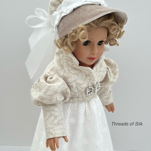 Puede incluir: Mu&ntilde;eca con un gorro beige con una cinta blanca y una chaqueta a juego sobre un vestido calado blanco. La chaqueta tiene un estampado floral y una broche de perlas. La mu&ntilde;eca tiene el pelo rubio rizado y lleva un vestido blanco.