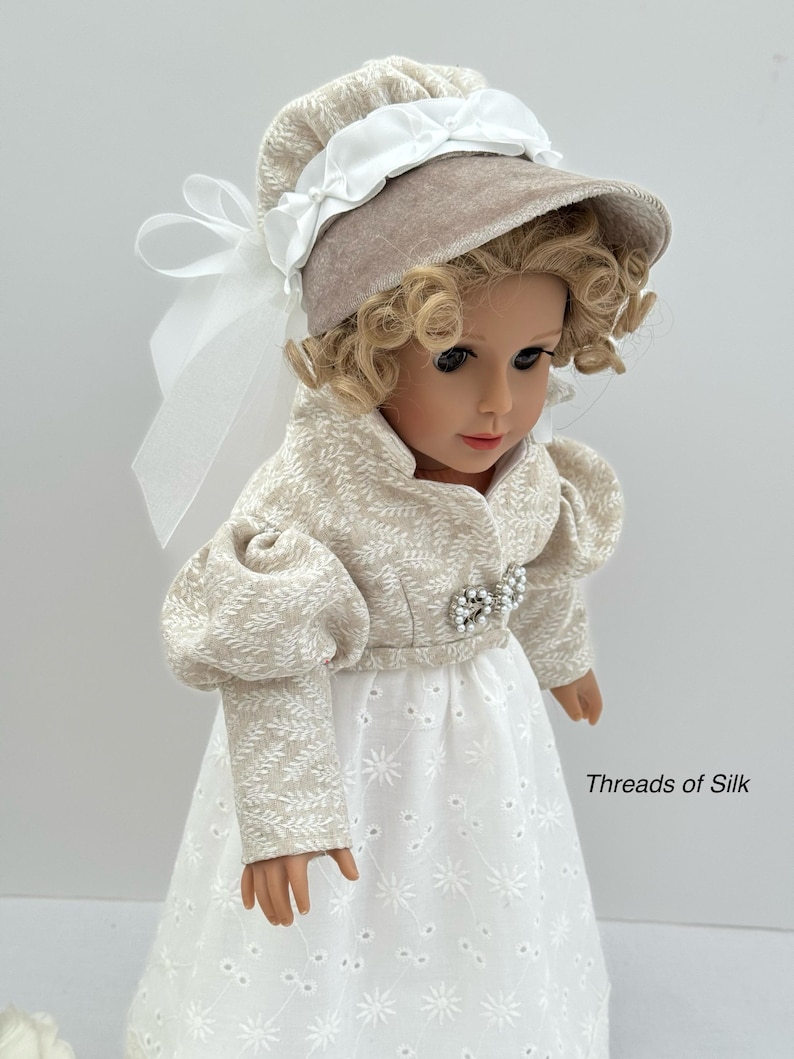 Puede incluir: Mu&ntilde;eca con un gorro beige con una cinta blanca y una chaqueta a juego sobre un vestido calado blanco. La chaqueta tiene un estampado floral y una broche de perlas. La mu&ntilde;eca tiene el pelo rubio rizado y lleva un vestido blanco.