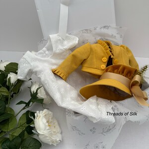 May include: A white eyelet dress, a mustard yellow jacket, and a matching hat with a ribbon are presented in an open white box. The box is lined with printed tissue paper. The text "Threads of Silk" is visible on the tissue paper.
