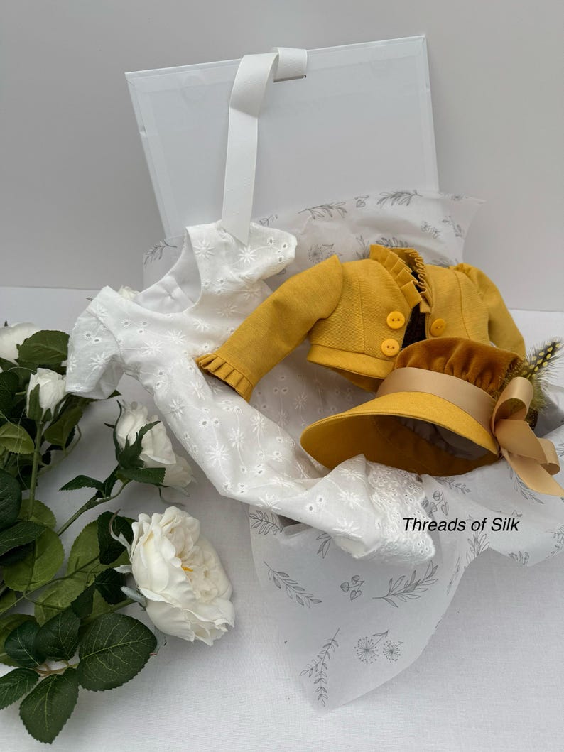 May include: A white eyelet dress, a mustard yellow jacket, and a matching hat with a ribbon are presented in an open white box. The box is lined with printed tissue paper. The text "Threads of Silk" is visible on the tissue paper.