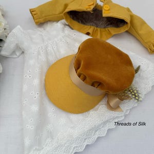 May include: A doll's outfit featuring a white eyelet dress, a mustard yellow jacket, and a matching hat with a brown velvet crown and ribbon. The outfit is displayed on a white surface. The text "Threads of Silk" is visible.