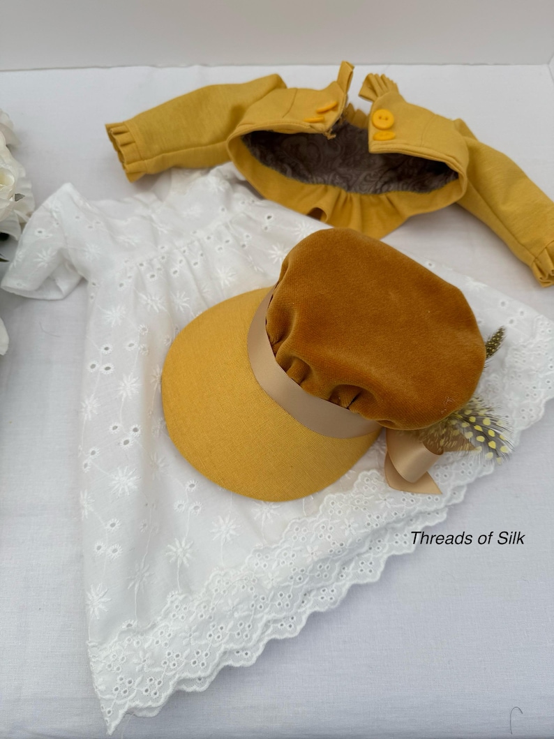 May include: A doll's outfit featuring a white eyelet dress, a mustard yellow jacket, and a matching hat with a brown velvet crown and ribbon. The outfit is displayed on a white surface. The text "Threads of Silk" is visible.