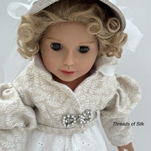 Puede incluir: Una mu&ntilde;eca vestida con un atuendo beige y blanco con un gorro a juego. El atuendo presenta un estampado floral y una broche de perlas. La mu&ntilde;eca tiene el pelo rubio rizado y lleva un vestido blanco. El texto "Threads of Silk" es visible.