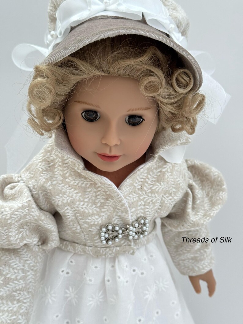 Puede incluir: Una mu&ntilde;eca vestida con un atuendo beige y blanco con un gorro a juego. El atuendo presenta un estampado floral y una broche de perlas. La mu&ntilde;eca tiene el pelo rubio rizado y lleva un vestido blanco. El texto "Threads of Silk" es visible.