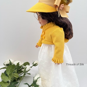 May include: A doll wearing a yellow hat with a wide brim and a matching yellow jacket with decorative buttons. The doll is also wearing a white eyelet skirt. The hat is adorned with a bow and feathers. The text "Threads of Silk" is visible.