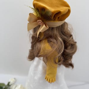 May include: Mustard yellow beret with a satin ribbon and a small flower accent. The beret is adorned with feathers and is set on a head of wavy, light brown hair. The beret is part of a vintage-inspired fashion ensemble.