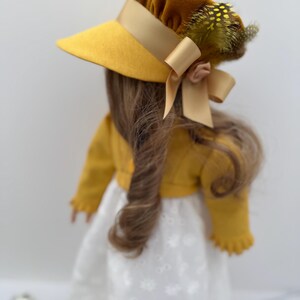 May include: A doll wearing a mustard yellow hat with a matching ribbon and a small bow. The hat is adorned with feathers. The doll is also wearing a mustard yellow jacket and a white dress with a floral pattern.