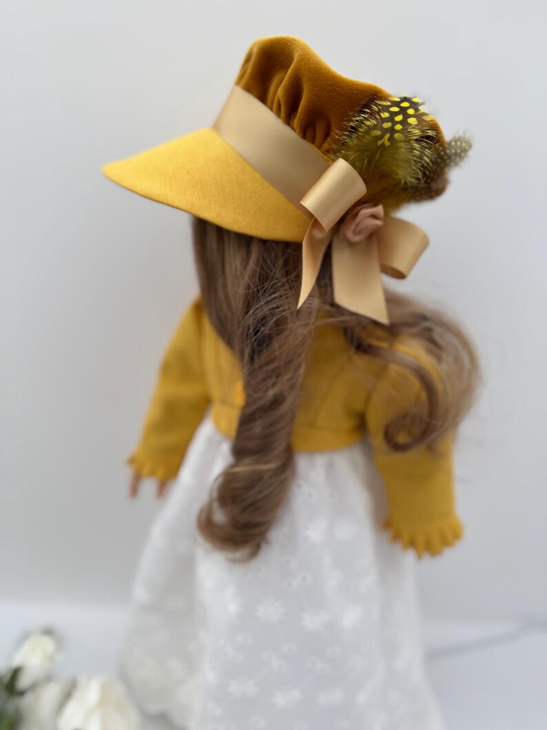 May include: A doll wearing a mustard yellow hat with a matching ribbon and a small bow. The hat is adorned with feathers. The doll is also wearing a mustard yellow jacket and a white dress with a floral pattern.