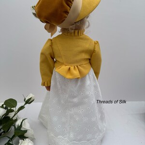 May include: A doll wearing a mustard yellow bonnet and jacket, paired with a white eyelet skirt. The outfit has a vintage aesthetic. The text "Threads of Silk" is visible on the skirt.