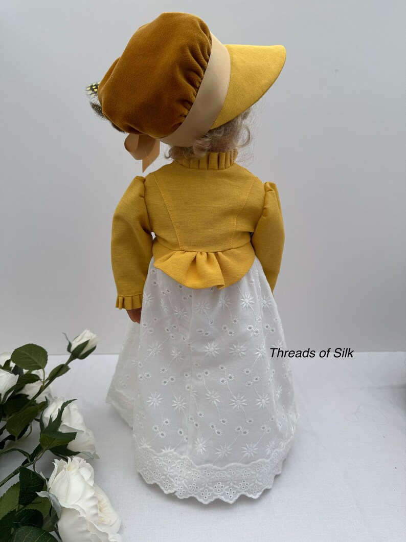 May include: A doll wearing a mustard yellow bonnet and jacket, paired with a white eyelet skirt. The outfit has a vintage aesthetic. The text "Threads of Silk" is visible on the skirt.