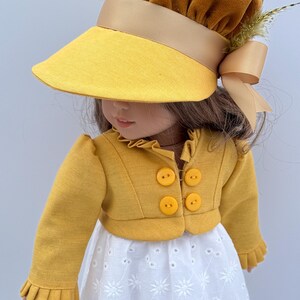 May include: A doll wearing a yellow sun visor with a brown velvet crown and a matching bow. The doll is also wearing a yellow jacket with four round buttons and a white eyelet dress. The outfit is a vintage style.