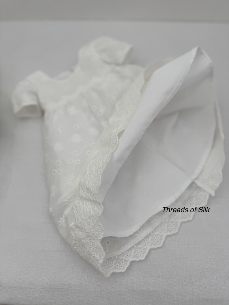 Puede incluir: Vestido blanco de ojal con mangas cortas y dobladillo de encaje festoneado. El vestido presenta un delicado bordado floral y el texto "Threads of Silk". Una prenda cl&aacute;sica y elegante.