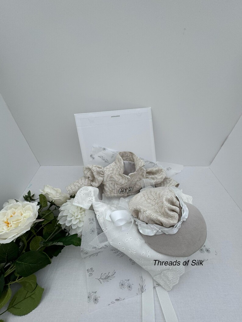 Puede incluir: Una chaqueta beige con estampado floral y un sombrero a juego con ala. El conjunto se acompa&ntilde;a de detalles florales blancos y una tela blanca con dise&ntilde;o floral. El texto "Threads of Silk" es visible.