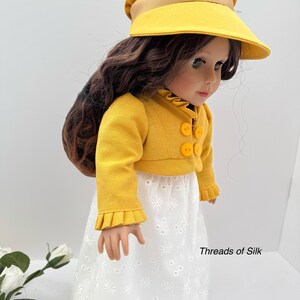 May include: A doll wearing a yellow jacket with round buttons, a white eyelet skirt, and a yellow visor hat with a brown brim. The doll has long brown hair. The text "Threads of Silk" is visible.
