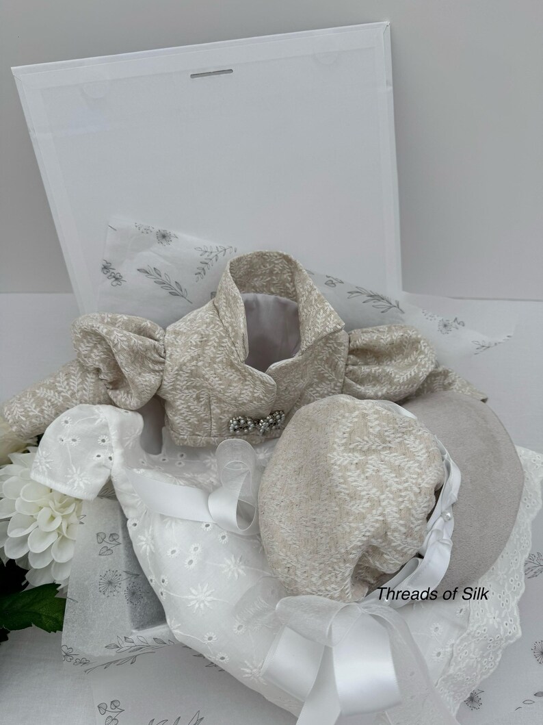 Puede incluir: Un conjunto de ropa de beb&eacute; beige y blanco, que incluye una chaqueta con mangas abullonadas, un gorro y un vestido blanco bordado. La chaqueta tiene un lazo decorativo. El conjunto se presenta en una caja blanca con papel de seda floral.