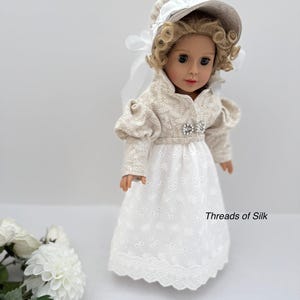 Puede incluir: Una mu&ntilde;eca vestida con un vestido crema de estilo Regencia, con un gorro y una chaqueta a juego. El vestido blanco presenta una falda bordada y una broche decorativa. El texto "Threads of Silk" es visible en el vestido.