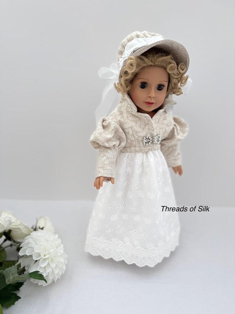 Puede incluir: Una mu&ntilde;eca vestida con un vestido crema de estilo Regencia, con un gorro y una chaqueta a juego. El vestido blanco presenta una falda bordada y una broche decorativa. El texto "Threads of Silk" es visible en el vestido.