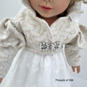 Puede incluir: Una mu&ntilde;eca vestida con una chaqueta color crema con estampado de hojas y un vestido calado blanco. La chaqueta tiene mangas abullonadas y una broche de perlas. El vestido tiene un dise&ntilde;o calado floral. El texto "Threads of Silk" es visible.