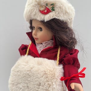 May include: A doll wearing a burgundy velvet coat and a matching hat with cream-coloured fur trim. The hat is decorated with a red stocking pin and a green sprig. The doll carries a cream-coloured fur purse with gold rope straps.