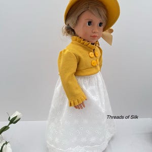 May include: A doll wearing a yellow hat and jacket with three yellow buttons. The doll is also wearing a white eyelet skirt. The text "Threads of Silk" is visible on the skirt.