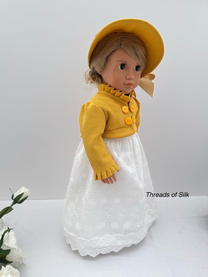 May include: A doll wearing a yellow hat and jacket with three yellow buttons. The doll is also wearing a white eyelet skirt. The text "Threads of Silk" is visible on the skirt.