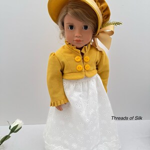 May include: A doll wearing a yellow jacket with three round buttons, a matching bonnet, and a white eyelet lace skirt. The doll has blonde hair and is wearing a yellow bonnet with a ribbon. The text "Threads of Silk" is visible on the skirt.