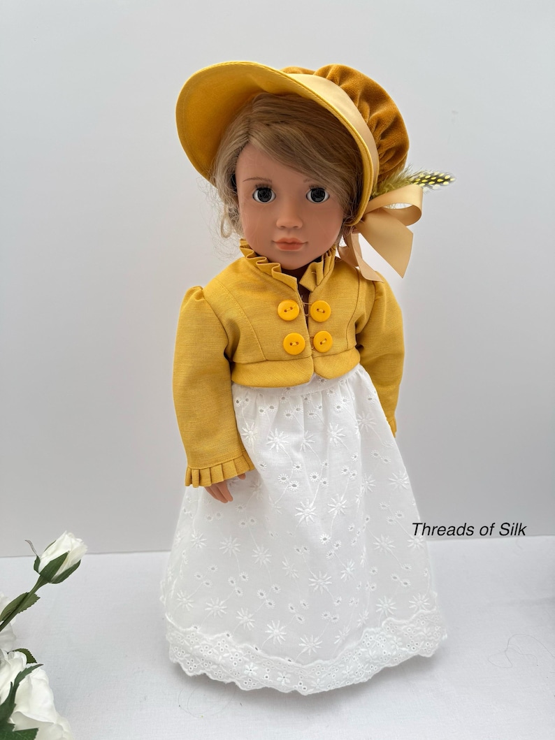 May include: A doll wearing a yellow jacket with three round buttons, a matching bonnet, and a white eyelet lace skirt. The doll has blonde hair and is wearing a yellow bonnet with a ribbon. The text "Threads of Silk" is visible on the skirt.