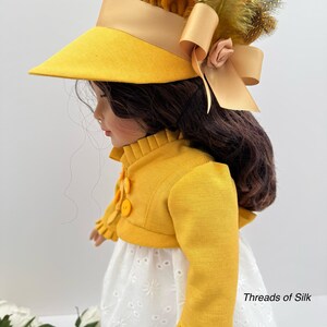 May include: A doll wearing a yellow hat with a wide brim, a matching jacket, and a white eyelet dress. The hat is adorned with a bow, feathers, and a small flower. The jacket has decorative buttons and ruffled cuffs. The text "Threads of Silk" is visible.